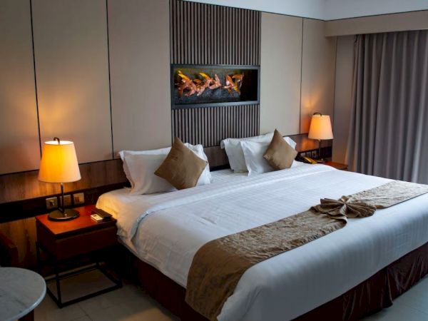 A hotel room with a neatly made bed, decorative pillows, lamps, and a small art piece above the bed, offering a cozy and inviting atmosphere.
