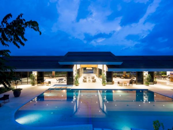 A modern building with a large, well-lit pool in the foreground, surrounded by seating areas and trees under a twilight sky.