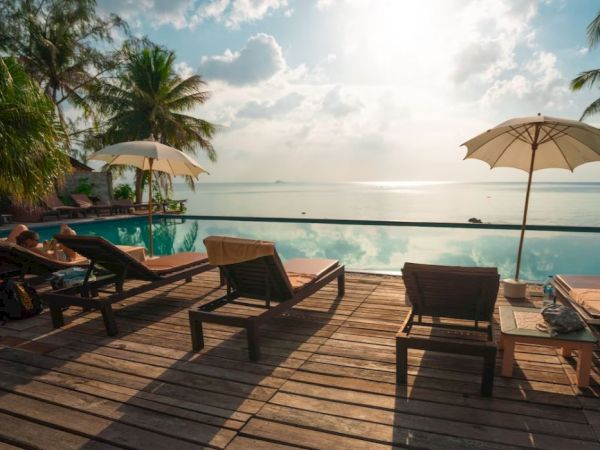 A serene seaside scene with lounge chairs, umbrellas, and a view of the horizon over calm waters and a pool on a partially shaded deck.