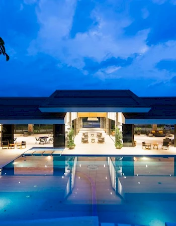 A modern building with a large, lit swimming pool in front, surrounded by seating, under a dramatic evening sky.
