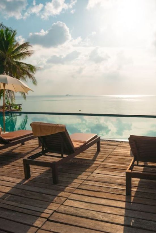 A serene tropical setting with lounge chairs by a pool, palm trees, and umbrellas, overlooking a calm body of water under a bright sky.