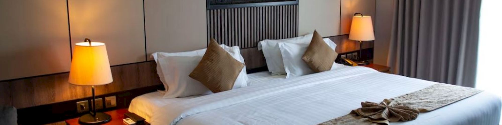 A neatly arranged hotel room with a large bed, decor pillows, bedside lamps, and a small table next to curtains in soft lighting.