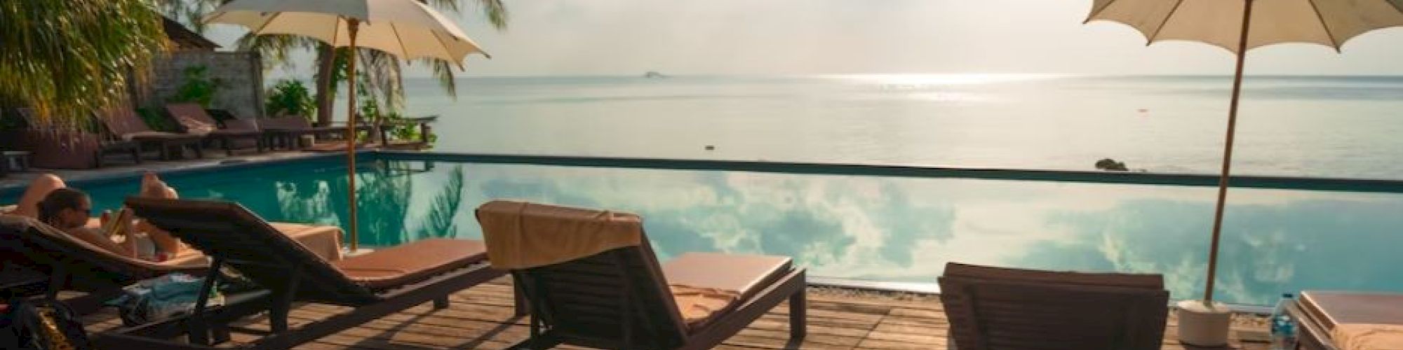 Poolside scene with lounge chairs, umbrellas, and a serene view of the water and sky. Sunlight reflects on the calm surface.