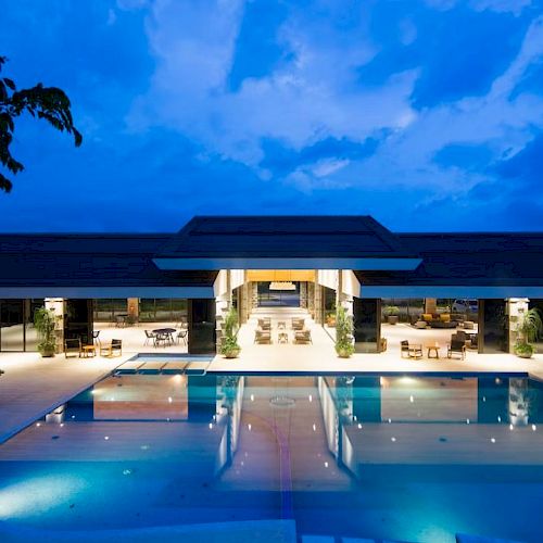A modern building with bright lights reflecting on a large swimming pool under a twilight sky.