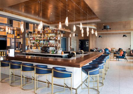 The image shows a stylish bar with blue stools, hanging lights, and a well-stocked counter against a modern, spacious background.
