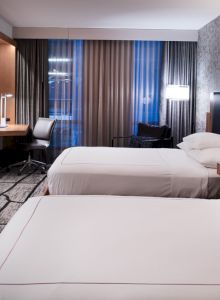 The image shows a modern hotel room with two beds, a TV, a desk, and a window with curtains, featuring a stylish, contemporary design.