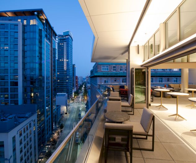 The image shows a modern cityscape with high-rise buildings and a rooftop terrace with seating, taken during dusk.