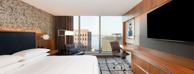 A modern hotel room with a large bed, wall-mounted TV, desk, and a window with a city view.