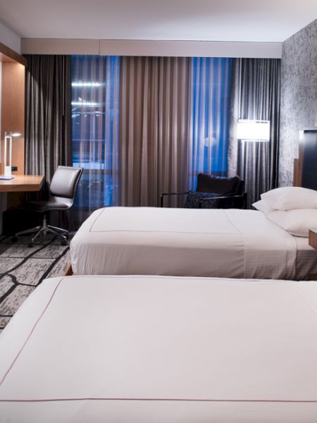 A modern hotel room with two beds, a wall-mounted TV, desk, and a large window with curtains, lit by calm, ambient lighting.