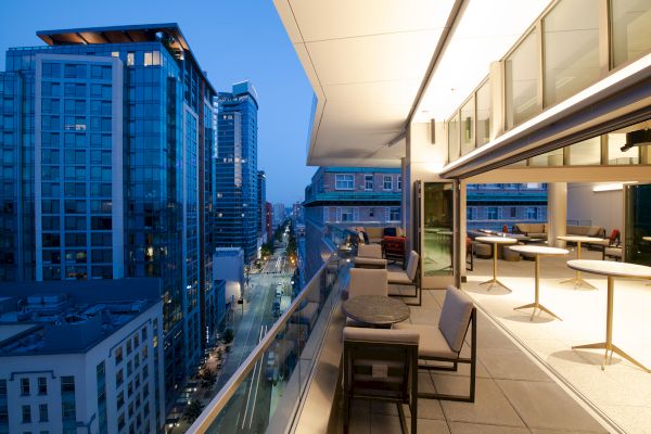 A modern cityscape at dusk shows a rooftop terrace with seating, buildings, and a street view, creating a vibrant urban atmosphere.