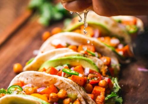 A hand squeezes lime over a row of colorful tacos filled with veggies and chickpeas, garnished with cilantro on a wooden board.