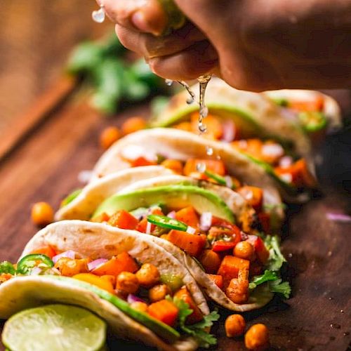 A hand squeezes lime over a row of colorful tacos filled with veggies and chickpeas, garnished with cilantro on a wooden board.