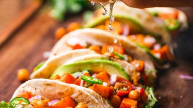 A hand squeezes lime over a row of colorful tacos filled with veggies and chickpeas, garnished with cilantro on a wooden board.