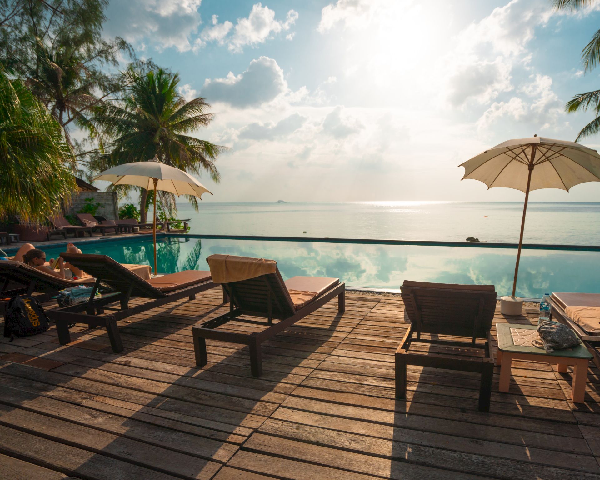 A serene poolside scene with lounge chairs, umbrellas, trees, and a calm body of water reflecting the bright sky.