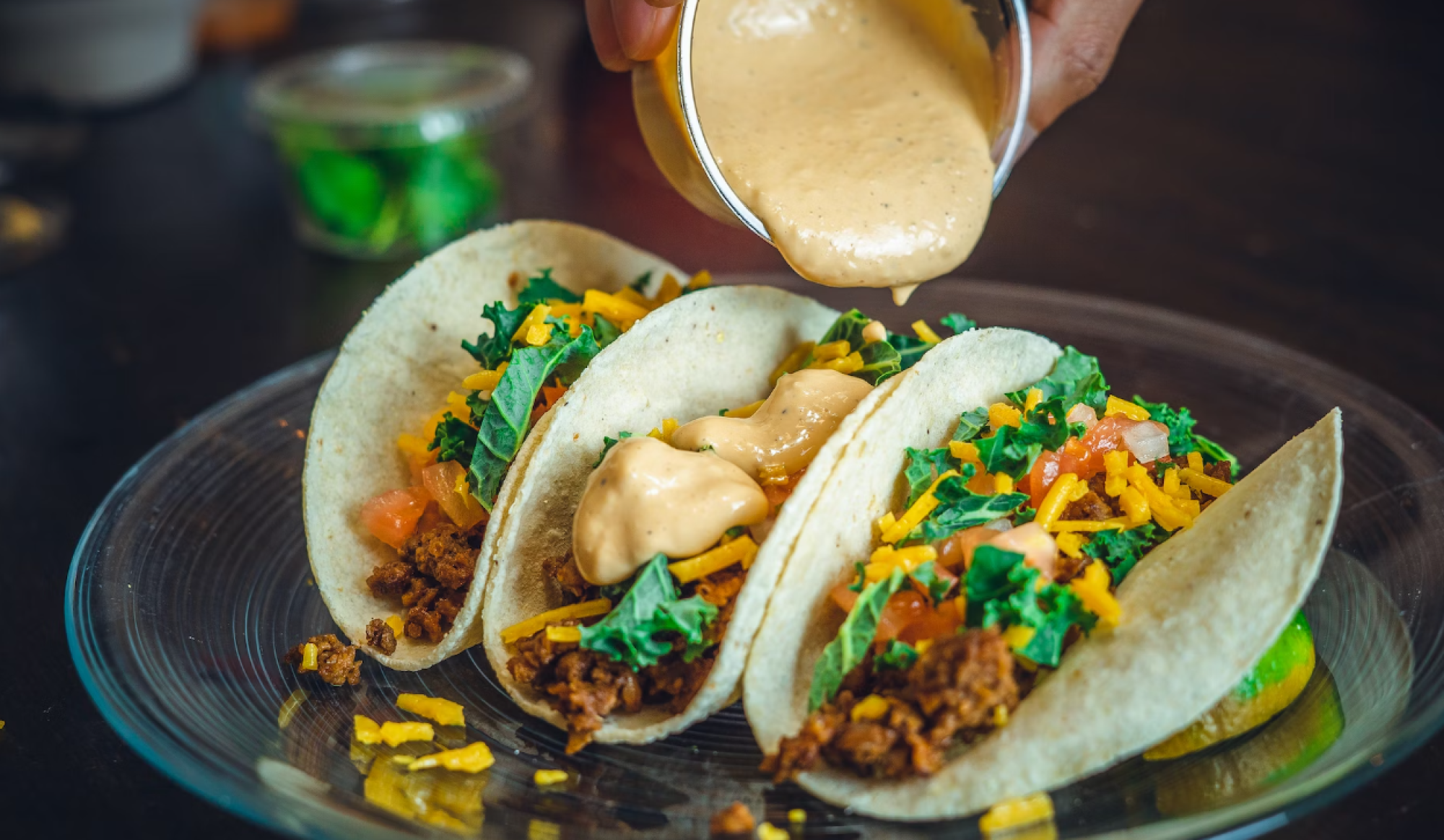 Three tacos on a plate being topped with creamy sauce, filled with meat, cheese, lettuce, and tomatoes, with a vibrant presentation.