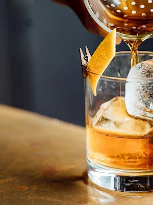 A cocktail is being poured into a glass with ice and an orange twist garnish, sitting on a bar counter.