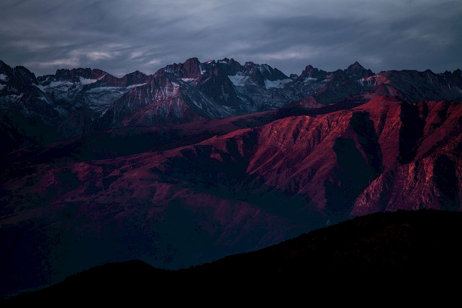 A mountain range bathed in deep red hues under a cloudy sky, creating a dramatic and moody landscape.