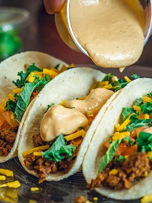 Soft tacos filled with ground meat, cheese, and greens, topped with a creamy sauce being poured over them on a plate.