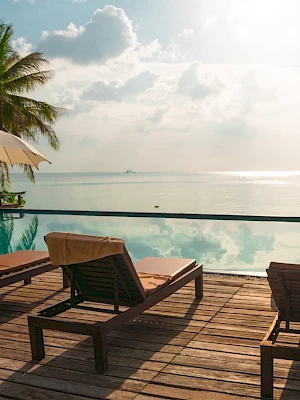 A serene poolside scene with lounge chairs, umbrellas, and palm trees overlooking a calm sea under a partly cloudy sky.