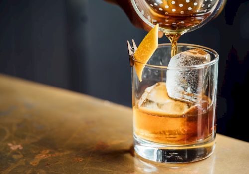 A cocktail being poured into a glass with ice and a lemon twist garnish, sitting on a wooden surface.