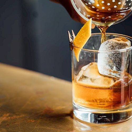 A cocktail being poured into a glass with ice and a lemon twist garnish, sitting on a wooden surface.