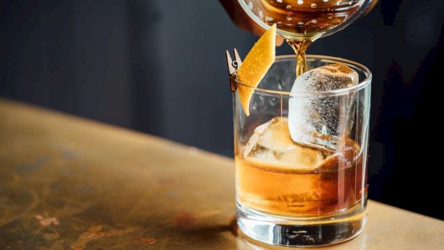 A cocktail being poured into a glass with ice and a lemon twist garnish, sitting on a wooden surface.
