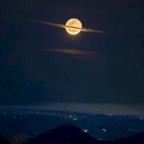 The image shows a full moon with clouds resembling Saturn's rings, above a landscape with distant city lights.