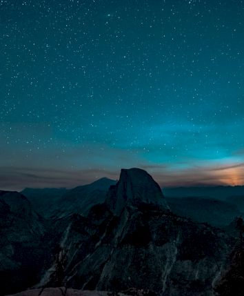 A starry night sky over a mountainous landscape with a glowing horizon and silhouettes of trees in the foreground.