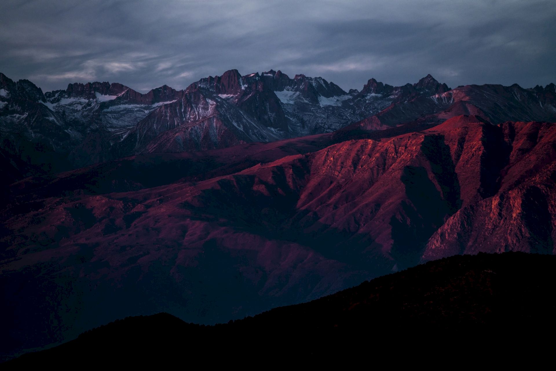 A mountain range at dusk, with deep red and purple hues from the sunset, under an overcast sky.