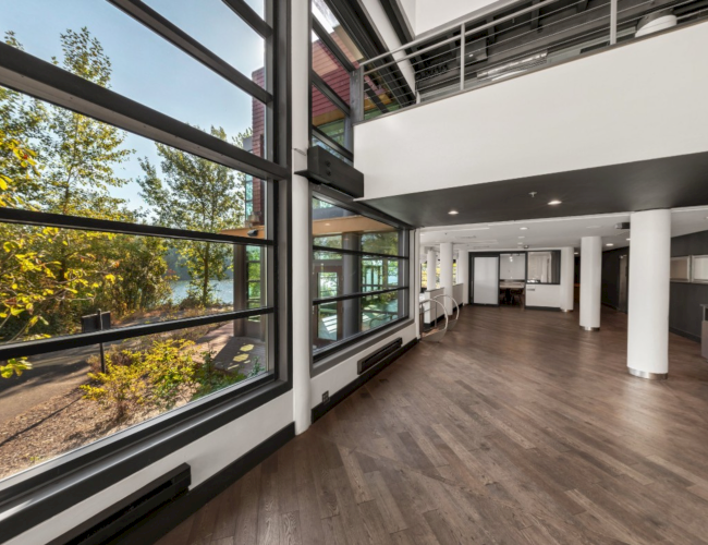 A modern indoor hallway with large glass windows, wood flooring, white columns, and a view of trees outside.