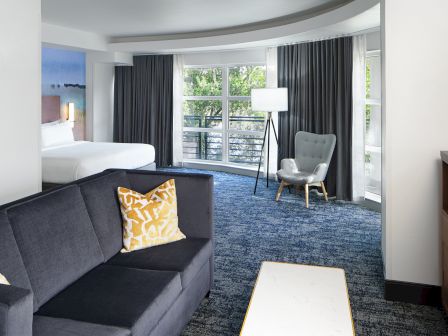 The image shows a modern hotel room with a bed, couch, coffee table, and large windows with curtains, creating a cozy atmosphere.