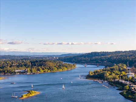 A panoramic river scene with sailboats, a small island, and tree-lined banks under a clear blue sky; a bridge spans the water in the distance.