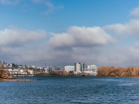 A cityscape with buildings under a cloudy blue sky is seen across a body of water, surrounded by trees with autumn foliage.