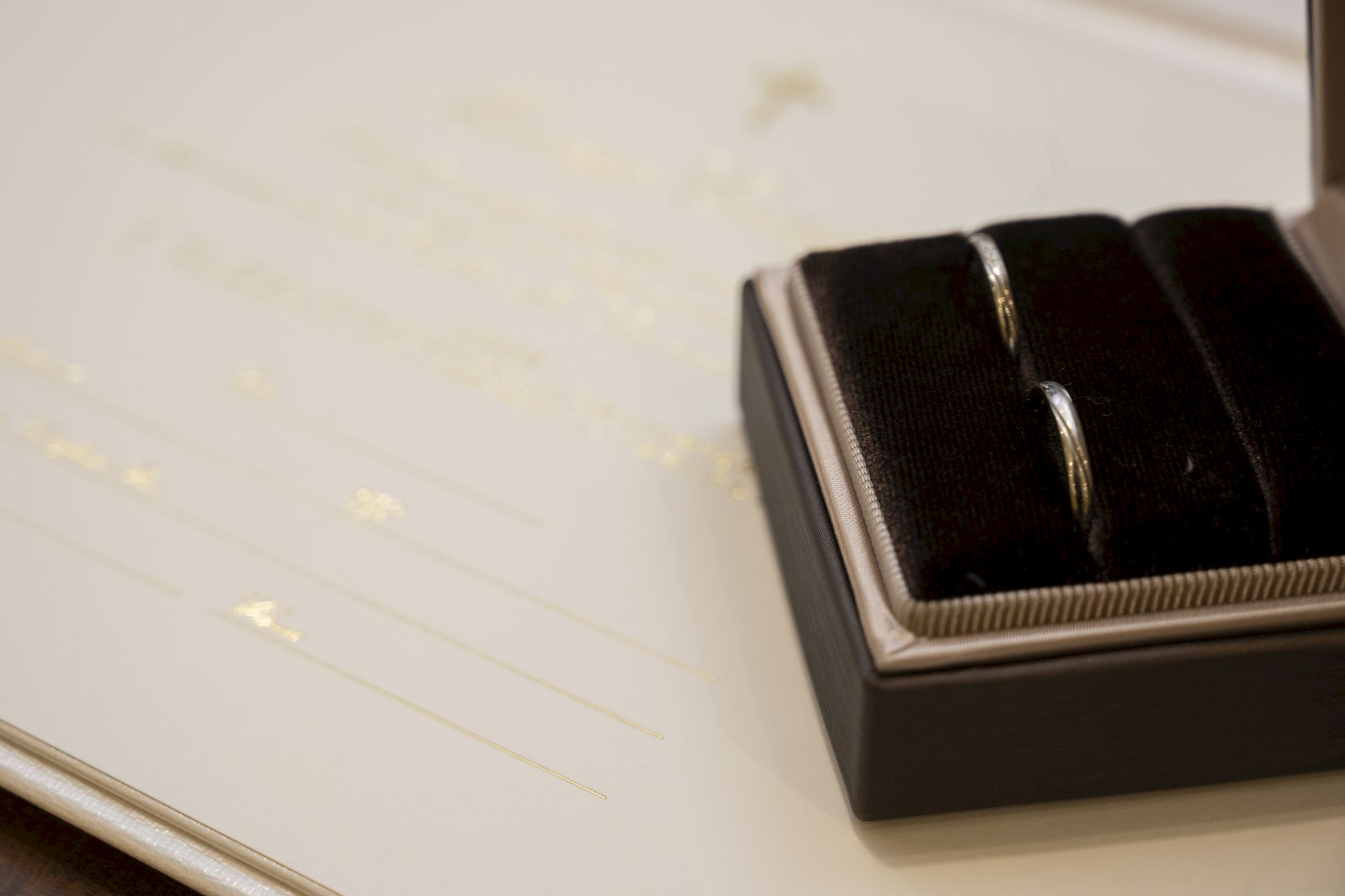 The image shows a pair of rings in a box placed on a document with gold lettering, possibly a wedding certificate.