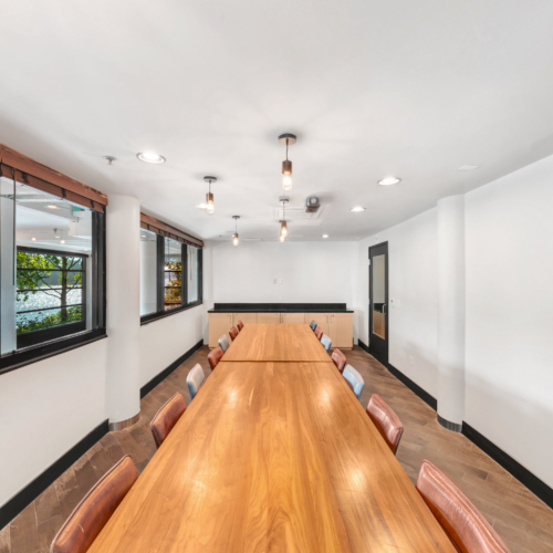 Bright conference room with a long wooden table, leather chairs, pendant lights, and large windows along one side.