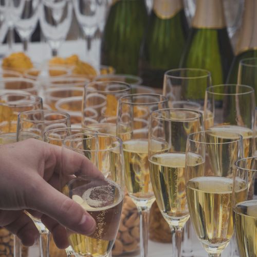 A hand places a glass among many filled with champagne; bottles and snacks are in the background, creating a celebratory setting.