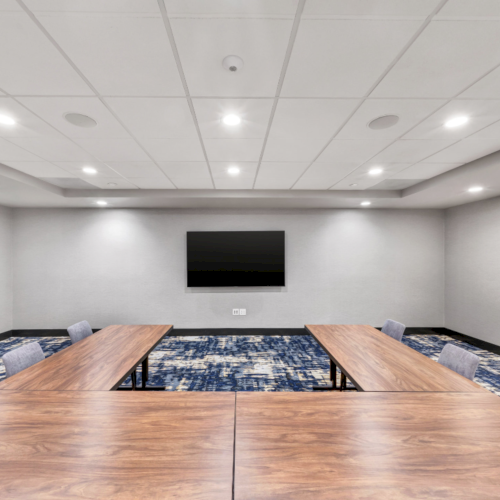 A modern conference room with two wooden tables, gray chairs, a mounted TV, blue patterned carpet, and recessed ceiling lights.