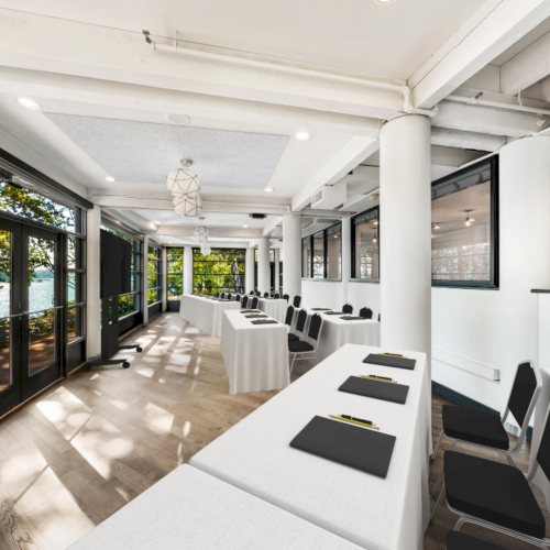 Bright modern meeting room with long white tables, black chairs, and large windows lining the side walls, under ceiling lights.