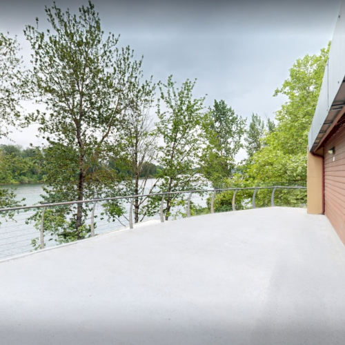 A lakeside house balcony with railings overlooks green trees and calm water under a cloudy sky.