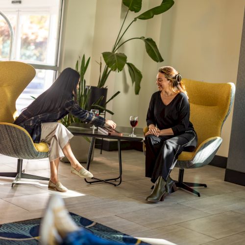 Two people chat casually in a bright lounge with yellow chairs, a plant, and a small coffee table between them, wine glasses on it.