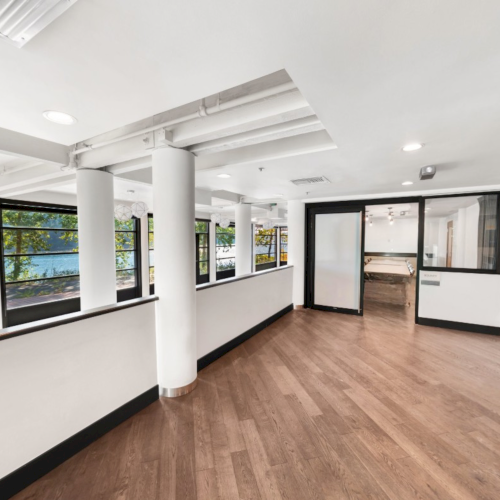 Bright modern office interior with wood floors, white columns, and large windows showing trees outside.