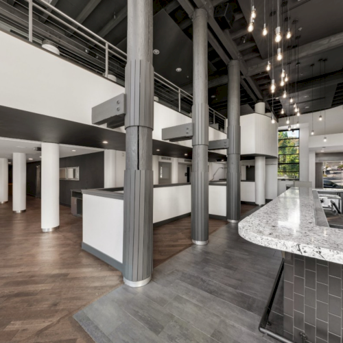 A modern open-plan office break area with a marble kitchen island, dark cabinets, pendant lights, and exposed concrete columns.