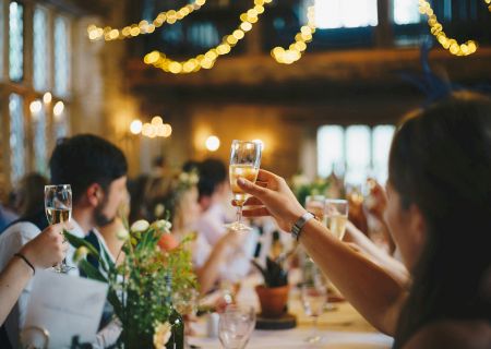 People are toasting with drinks at a festive gathering in a warmly lit, elegantly decorated space with string lights overhead.
