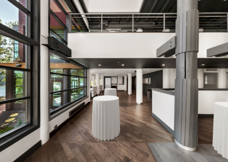 A modern interior hall with large windows, white-covered tables, tall columns, and a sleek bar-style counter in a bright venue.