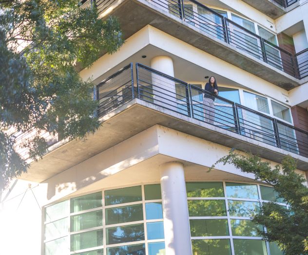 A person leaning on the balcony railing of a modern multi-story building, with trees nearby and sunlight shining on the corner.