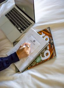 A person writes in a notebook on a bed beside a laptop and two comics; the scene suggests studying or journaling in a cozy setup.
