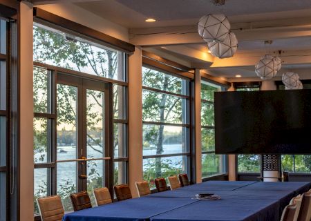 A long conference room with a blue-draped table, surrounded by beige chairs, large windows, and modern geometric pendant lights.