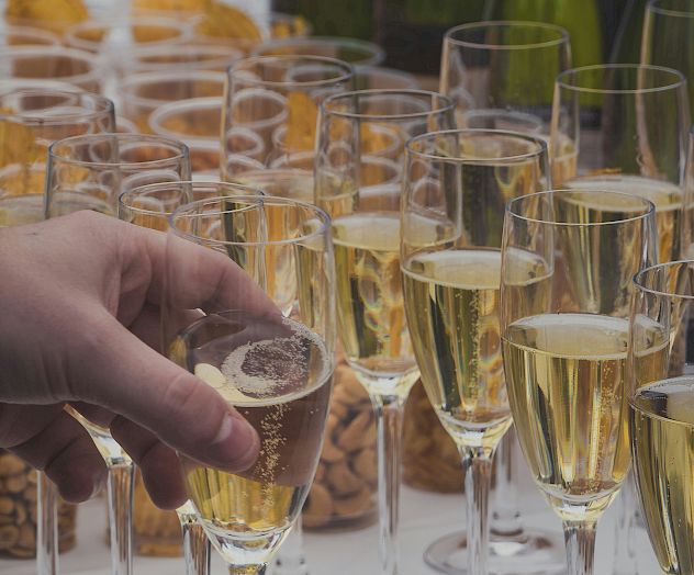 A hand holds a champagne flute among many filled glasses on a table, with bottles and snacks in the background.