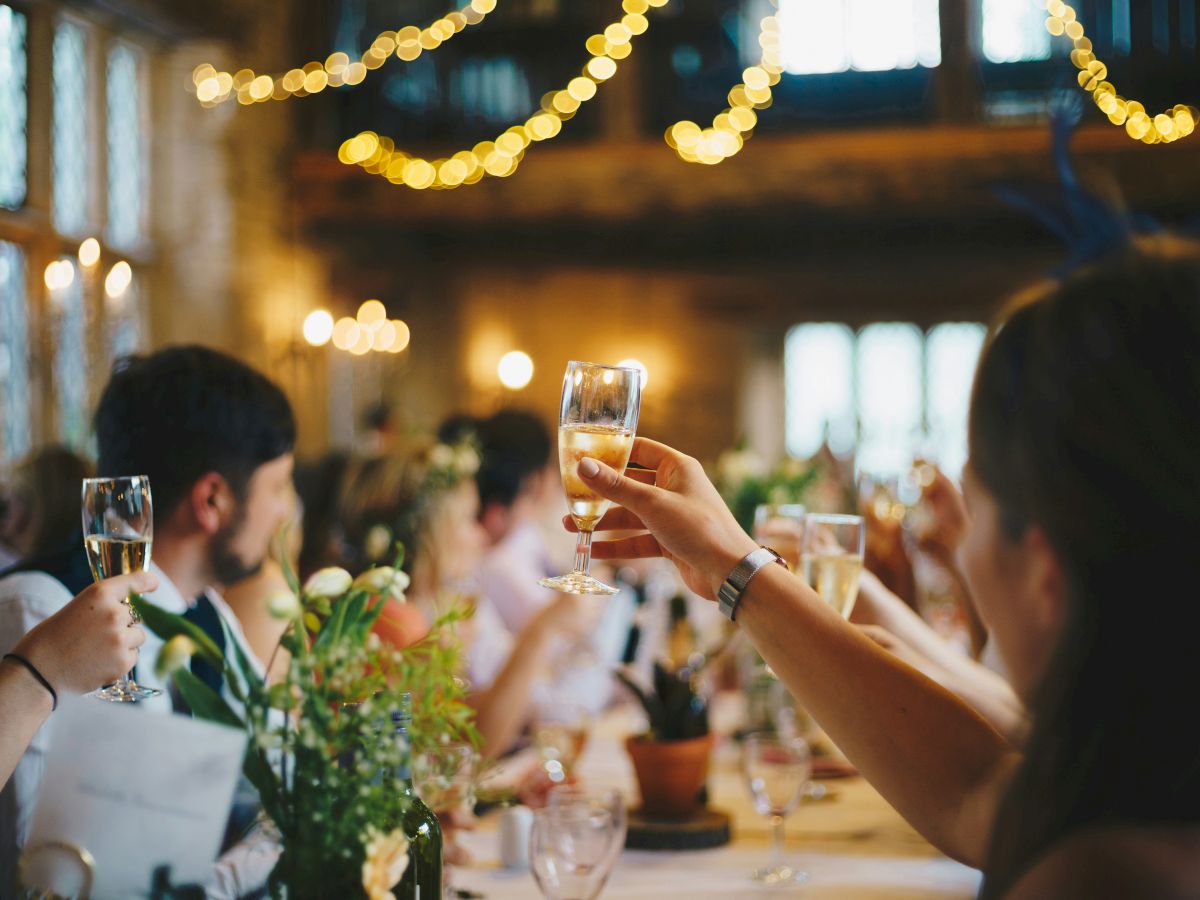 People raising glasses in a celebration, surrounded by a decorated venue with string lights and floral arrangements.