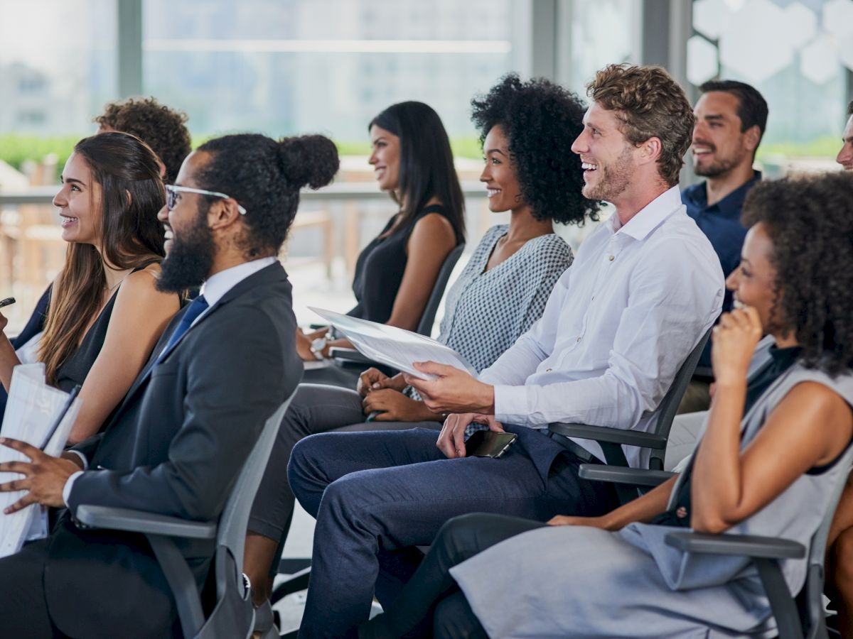 People seated in a room, appearing engaged, as if attending a presentation or seminar, with pens and papers in hands.